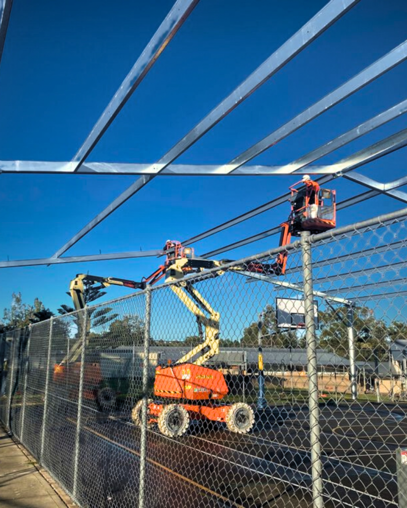 Rigging team installing a steel COLA structure over basketball courts at Kariong High School using elevated work platforms.