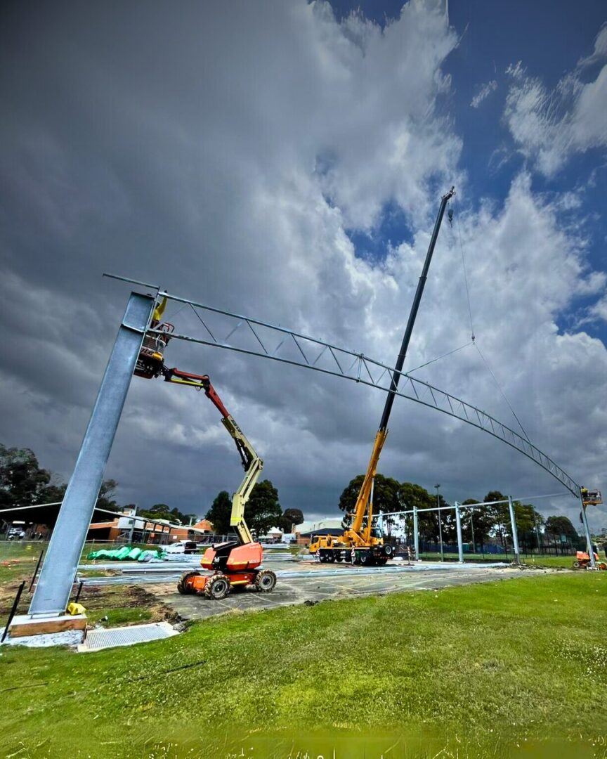 Structural steel erection for a COLA basketball court at Ambarvale High School, with cranes and boom lifts positioning large steel trusses.