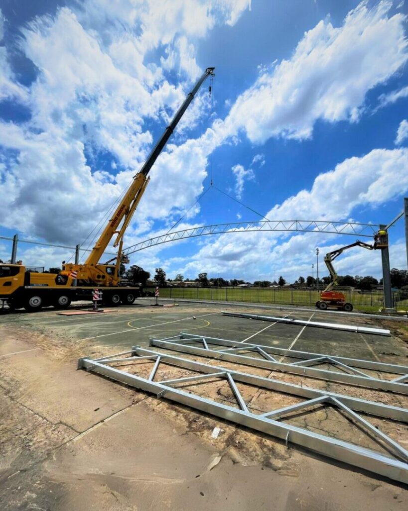 Crane lifting a steel roof truss during installation of a covered outdoor learning area (COLA) at a school facility.