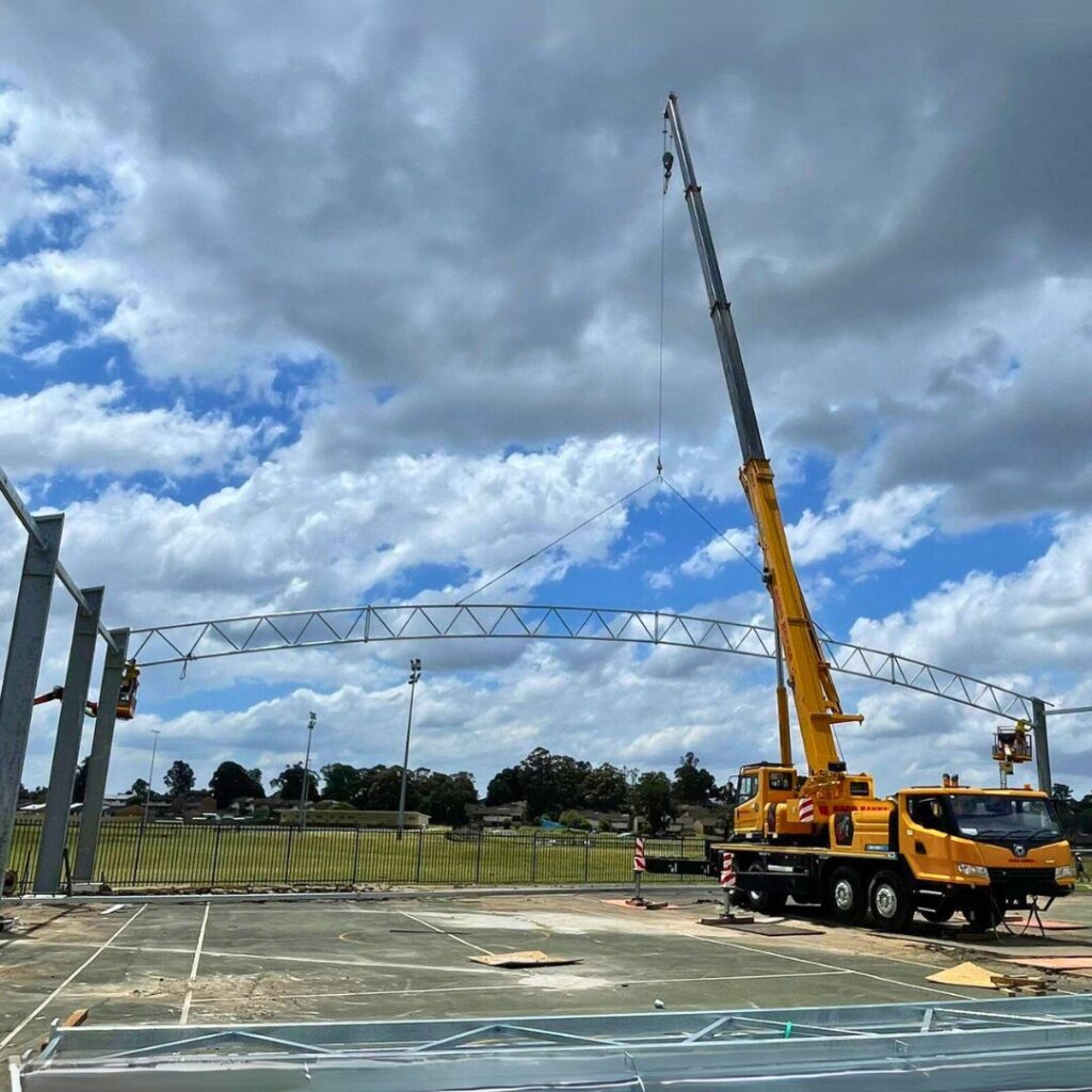 Yellow mobile crane lifting a steel truss beam for a COLA shelter at Ambervale High School’s outdoor basketball court.
